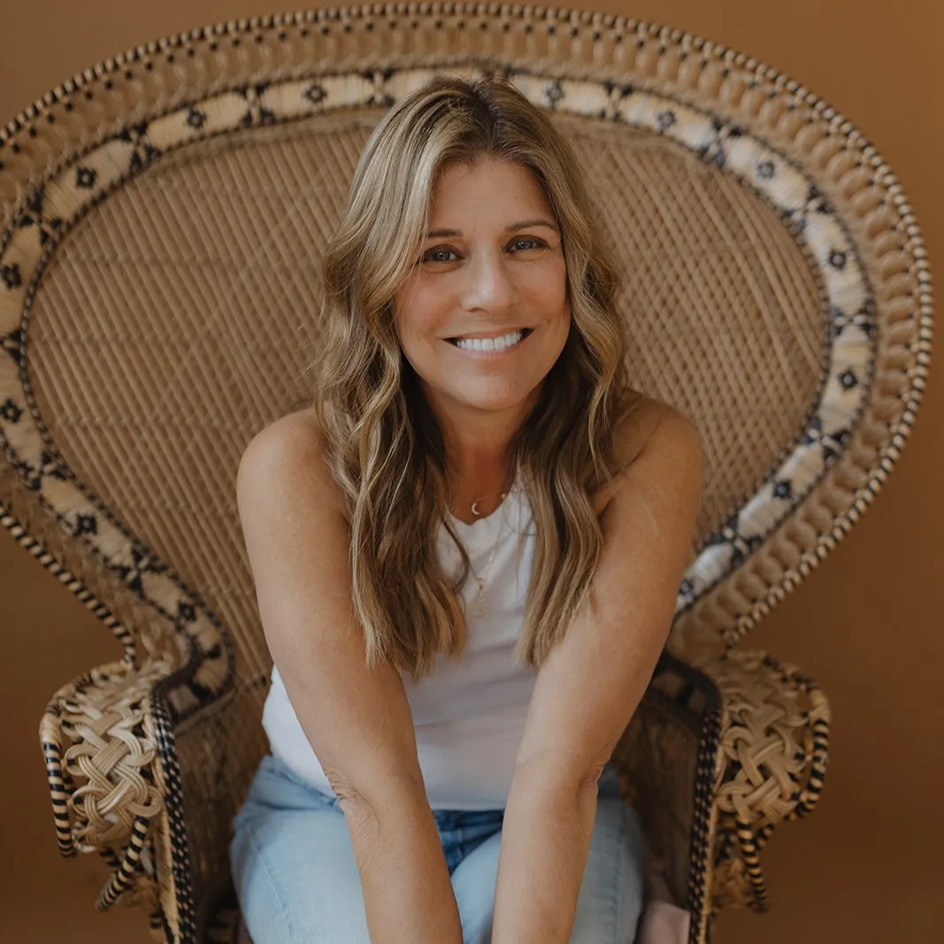 Smiling woman with wavy blonde hair sitting on an ornate woven chair against a brown background.